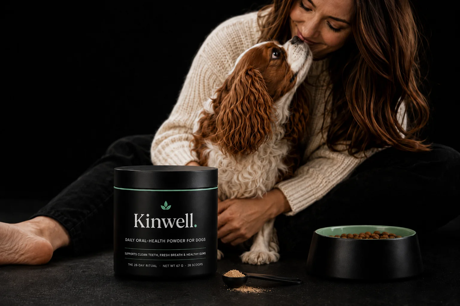 Owner sitting on a dark floor with her Cavalier King Charles Spaniel, a jar of Kinwell daily oral-health powder in the foreground
